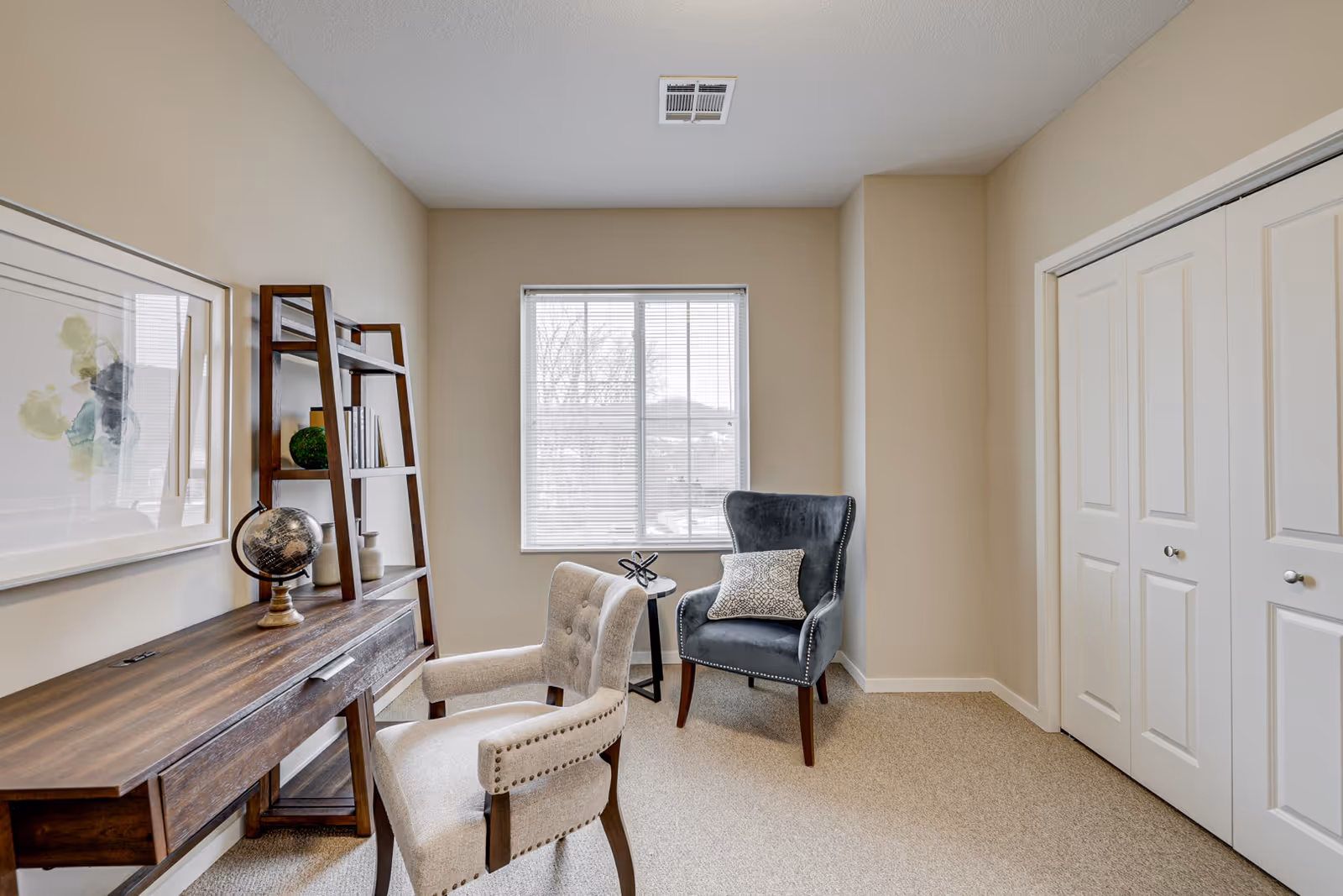 A small, cozy room with beige walls and carpet. There is a wooden desk with a globe and decorative items on it, accompanied by a beige upholstered chair with wooden legs. Next to the desk is a wooden ladder-style bookshelf with books and decor. A blue upholstered armchair with a patterned pillow sits near a window with blinds. On the right side, there are white double closet doors.