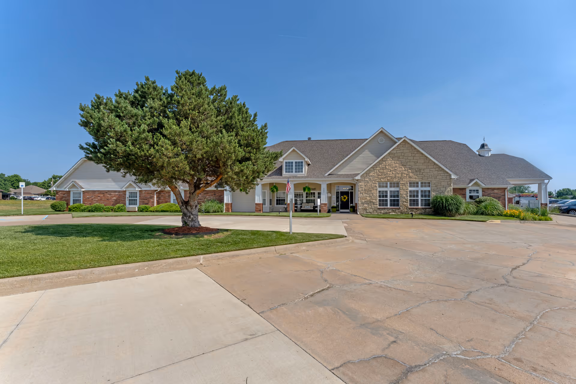 Exterior view of a single-story senior living facility building with a combination of brick and stone facade, a large tree in front on a grassy area, and a paved parking lot under a clear blue sky.