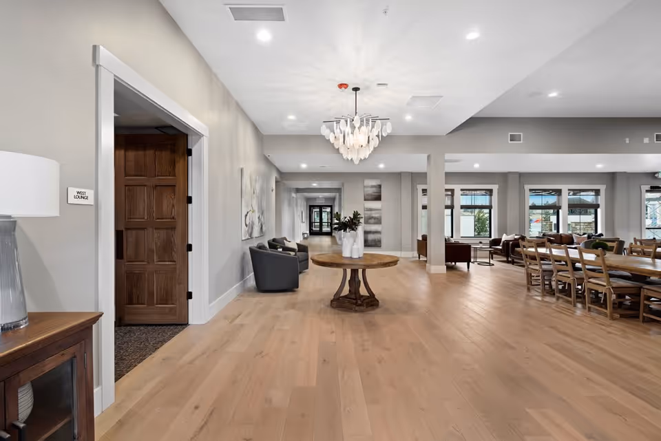 Spacious and well-lit interior lounge area with wooden flooring, a round wooden table with white vases in the center, several chairs and sofas arranged near large windows, and a chandelier hanging from the ceiling. A wooden door labeled 'West Lounge' is visible on the left side.