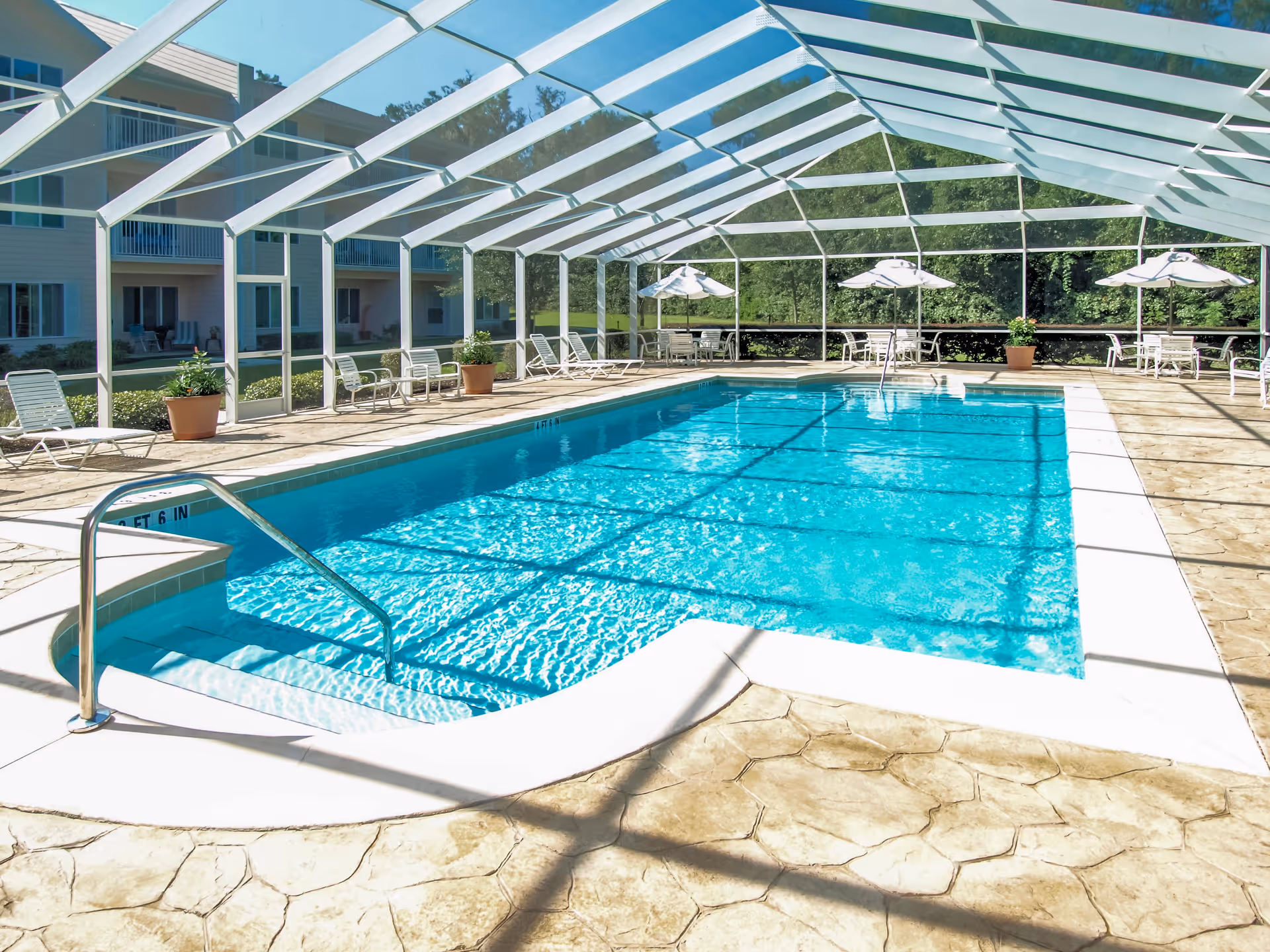 A rectangular swimming pool inside a screened enclosure with lounge chairs, umbrellas, and an adjacent multi-story building.