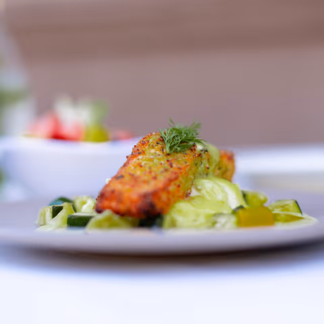 Close-up of a plated dish featuring a seasoned and cooked piece of fish garnished with a sprig of dill, served on a bed of vegetables with a creamy sauce.