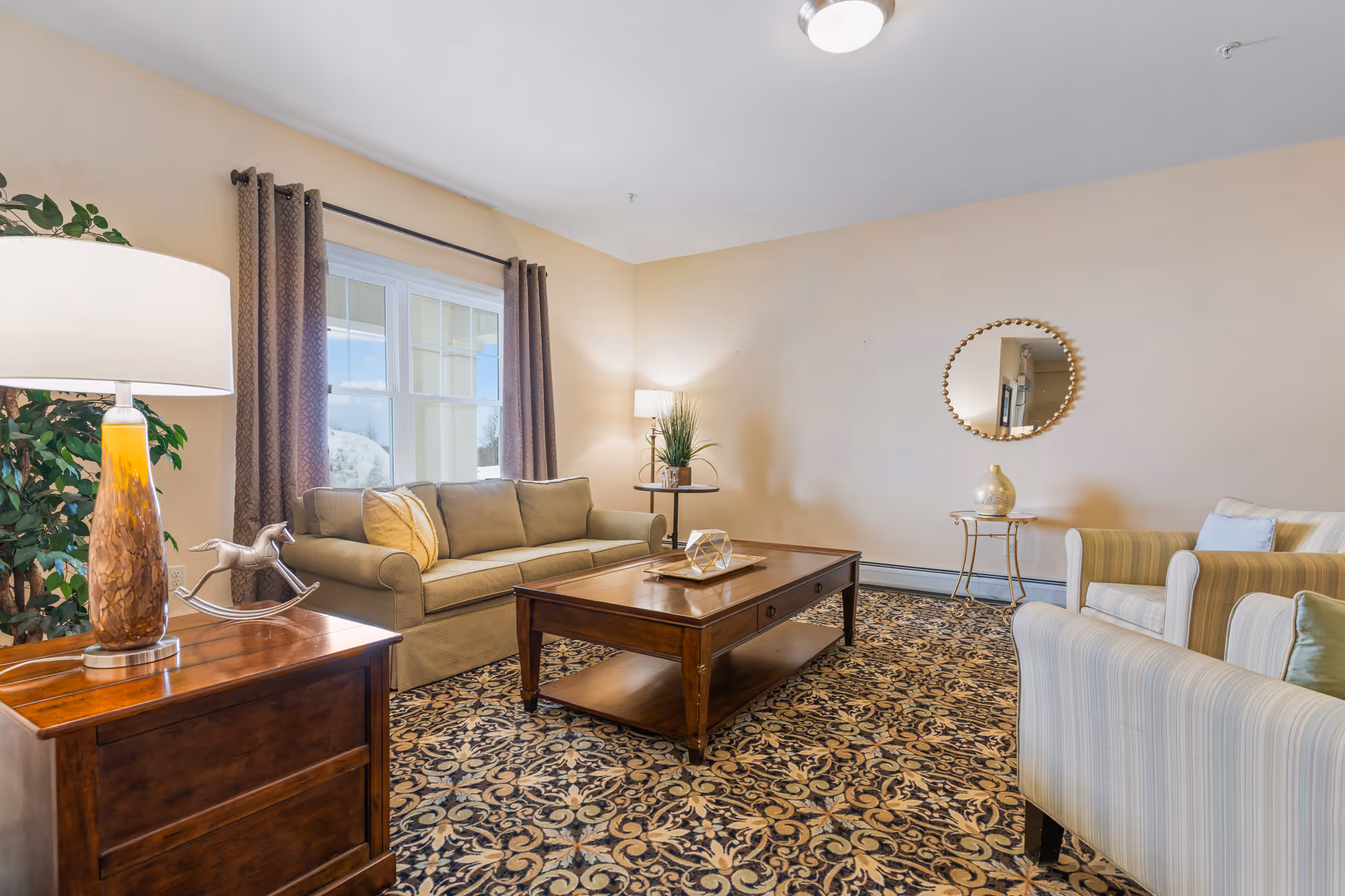 A cozy living room in a senior living facility with a beige sofa, two striped armchairs, a wooden coffee table, a side table with a lamp, a decorative mirror on the wall, and a patterned carpet. A large window with brown curtains lets in natural light.