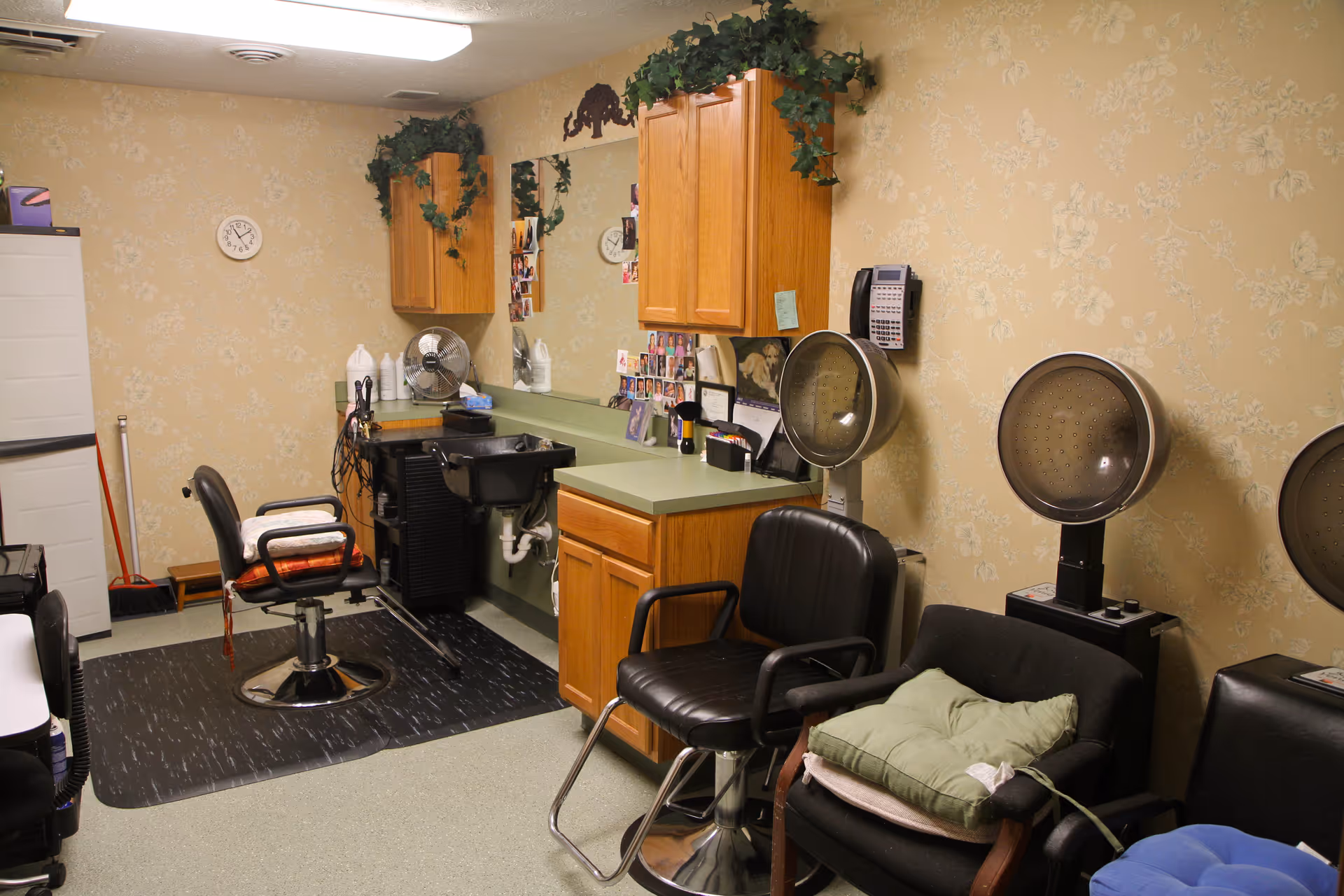 Small salon room with styling chairs, hooded hair dryers, a shampoo sink, mirror and wooden cabinets.