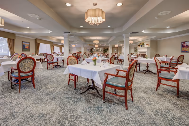 A spacious dining room with multiple tables covered in white tablecloths, each set with napkins and small flower arrangements. The room features patterned carpet, elegant wooden chairs with upholstered backs and seats, soft lighting from ceiling fixtures, and large windows with curtains allowing natural light. The walls are decorated with framed artwork and there is a fireplace on the right side.