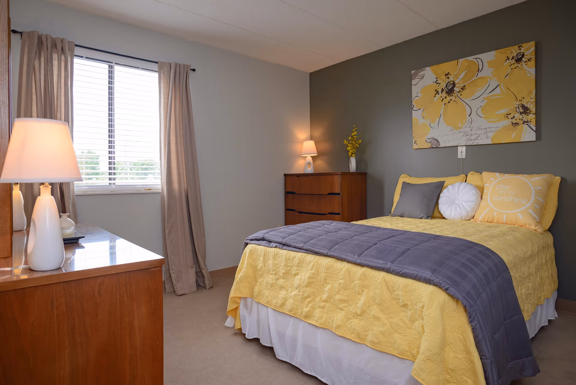 A cozy bedroom with a bed dressed in yellow and gray bedding, including decorative pillows. There is a wooden dresser with a lamp and a vase on it, and another dresser with a lamp and a yellow flower vase in the corner. A window with beige curtains allows natural light into the room. A large floral painting with yellow flowers hangs on the gray accent wall behind the bed.