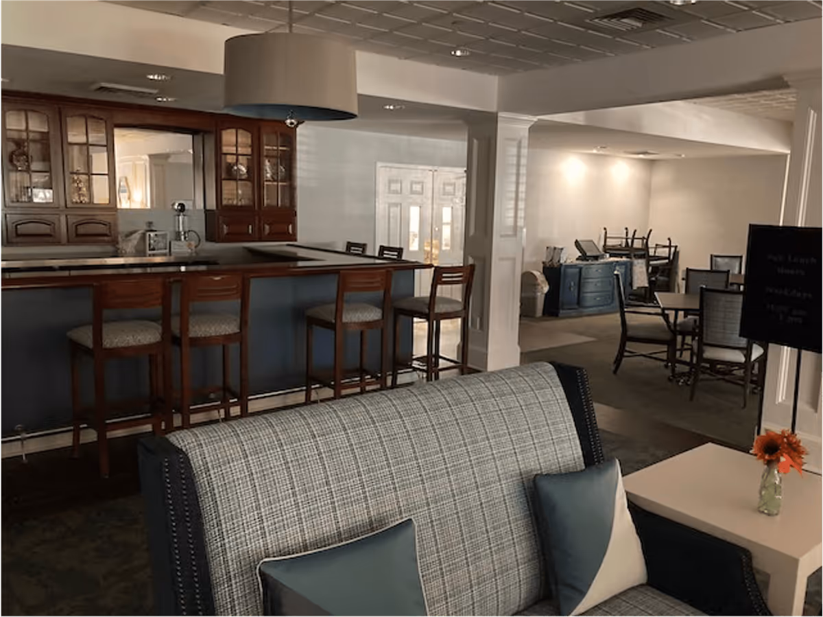 Interior view of a retirement community common area featuring a bar with four wooden bar stools, a cushioned sofa with two pillows, a small table with a vase holding orange flowers, and a dining area with tables and chairs in the background.