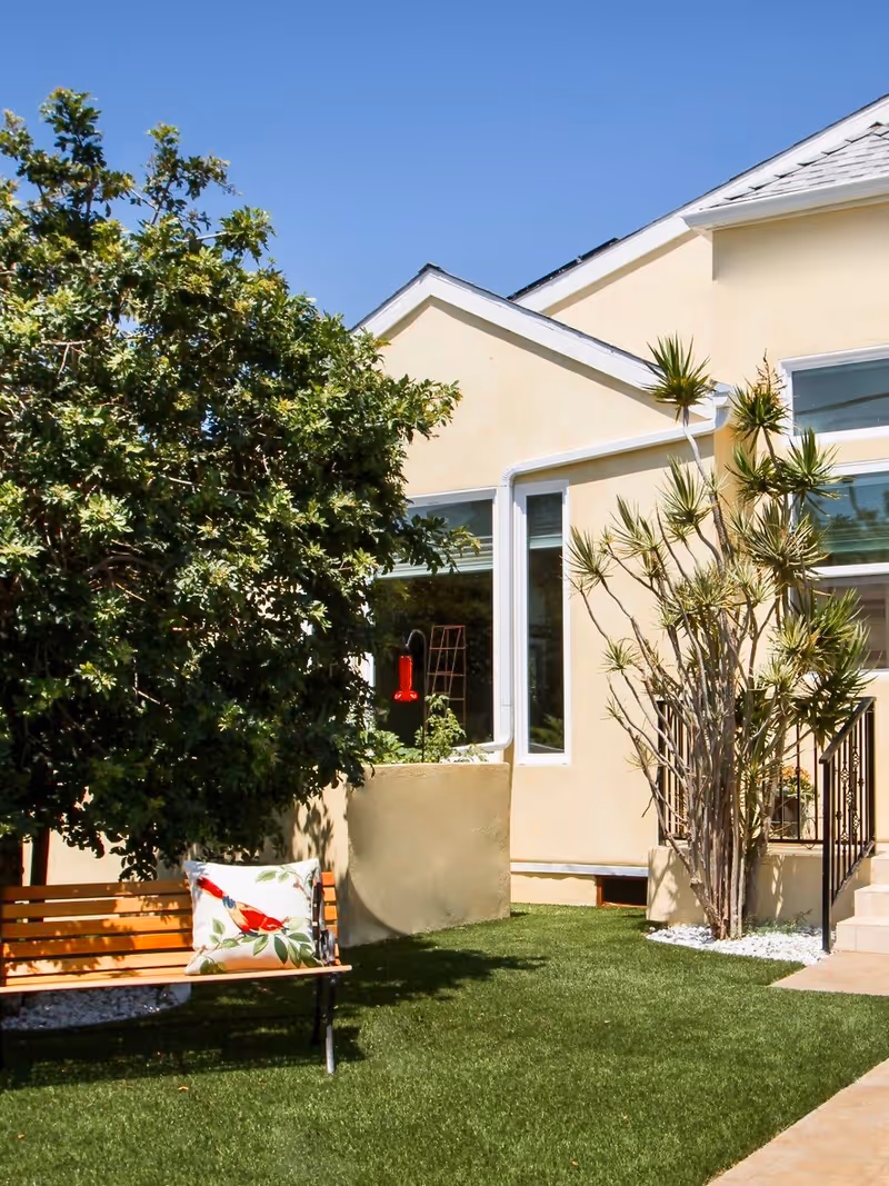 Outdoor garden area with a wooden bench featuring a decorative pillow with a bird design, surrounded by green grass, trees, and plants next to a light yellow building under a clear blue sky.