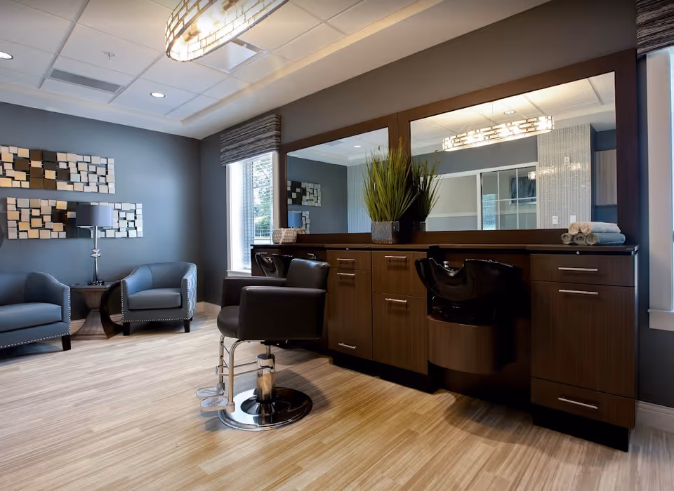 A modern salon area in a senior living facility featuring two black salon chairs in front of a large mirror with wooden cabinetry below. To the left, there are two blue armchairs with a small round table and a lamp between them. The walls are painted gray with decorative wall art, and there is a window letting in natural light.