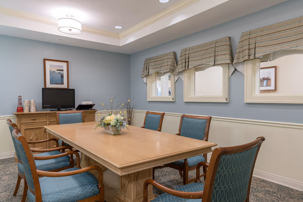 A small meeting or dining room with a rectangular wooden table surrounded by six wooden chairs with blue patterned cushions. On the table is a floral centerpiece. The walls are painted light blue on the upper half and cream on the lower half, with three windows covered by striped valances. A wooden sideboard against the wall holds a small TV, a framed picture, and decorative items. The ceiling has a recessed light fixture.