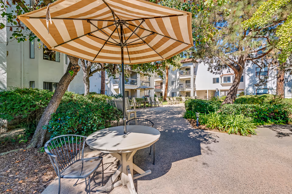 Outdoor seating area at a senior living facility with a round table and two metal chairs under a large striped umbrella. The area is surrounded by trees, bushes, and a paved walkway leading to a multi-story white building with balconies.