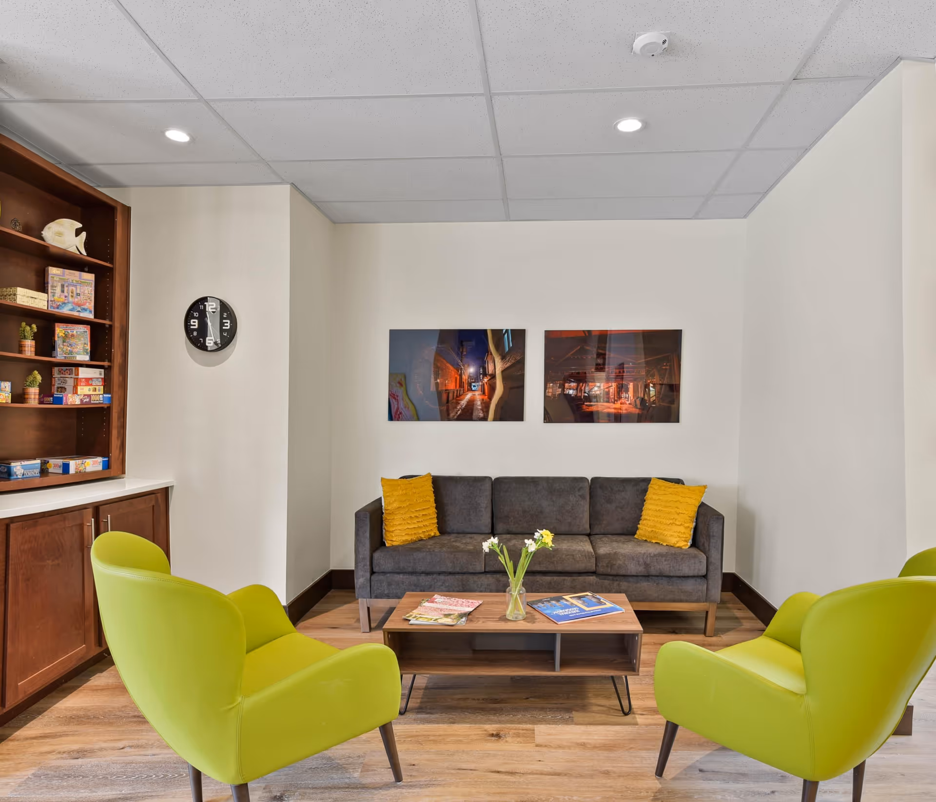 A cozy senior living lounge area with a dark gray sofa adorned with two yellow pillows, two bright green armchairs, and a wooden coffee table with a vase of flowers and magazines. There are two framed pictures on the wall above the sofa, a wall clock, and a wooden shelving unit filled with board games and decorative items.