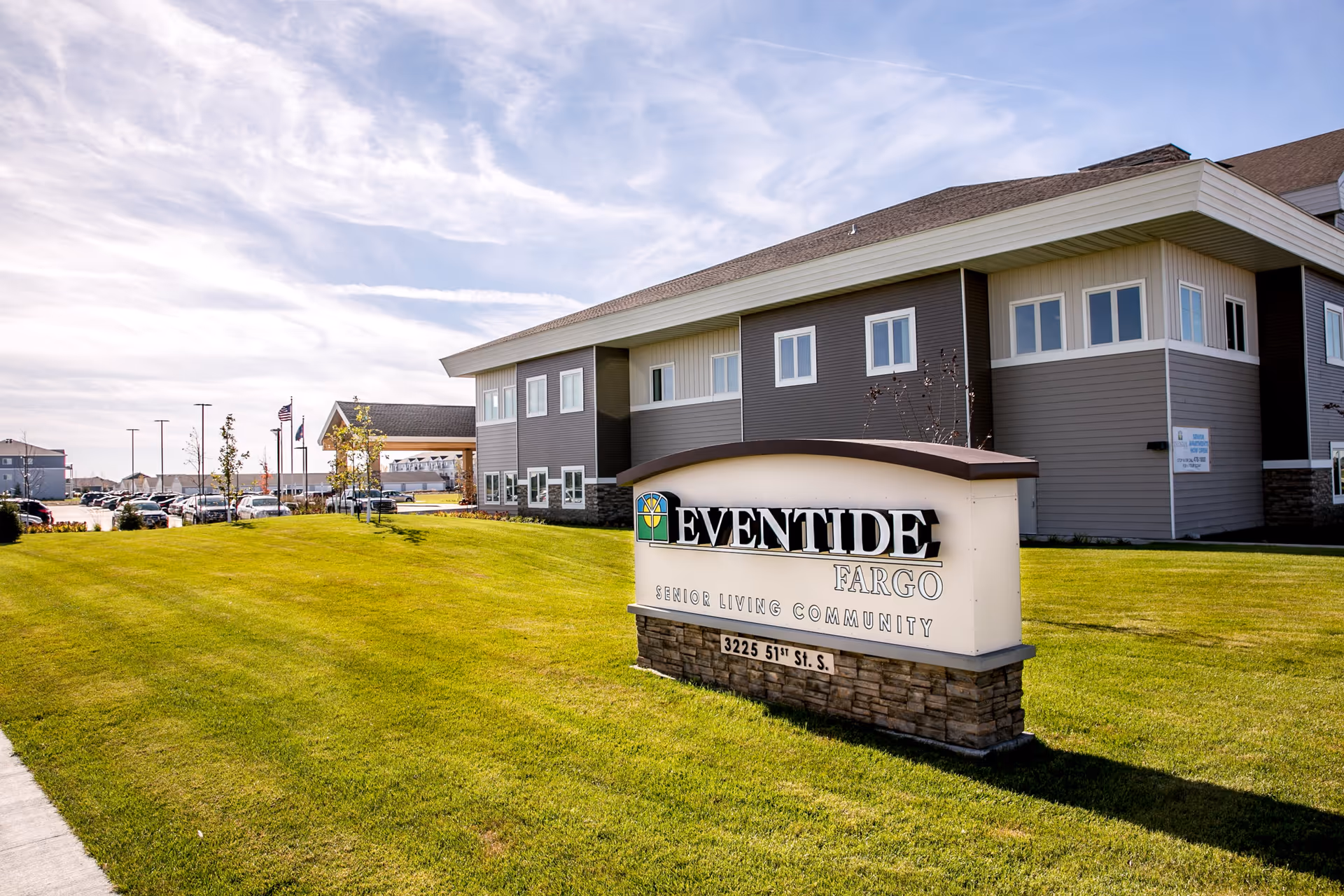 Exterior view of Eventide Fargo senior living community building with a large sign in the foreground displaying the facility name and address. The building is two stories with multiple windows and surrounded by a well-maintained green lawn under a partly cloudy sky.
