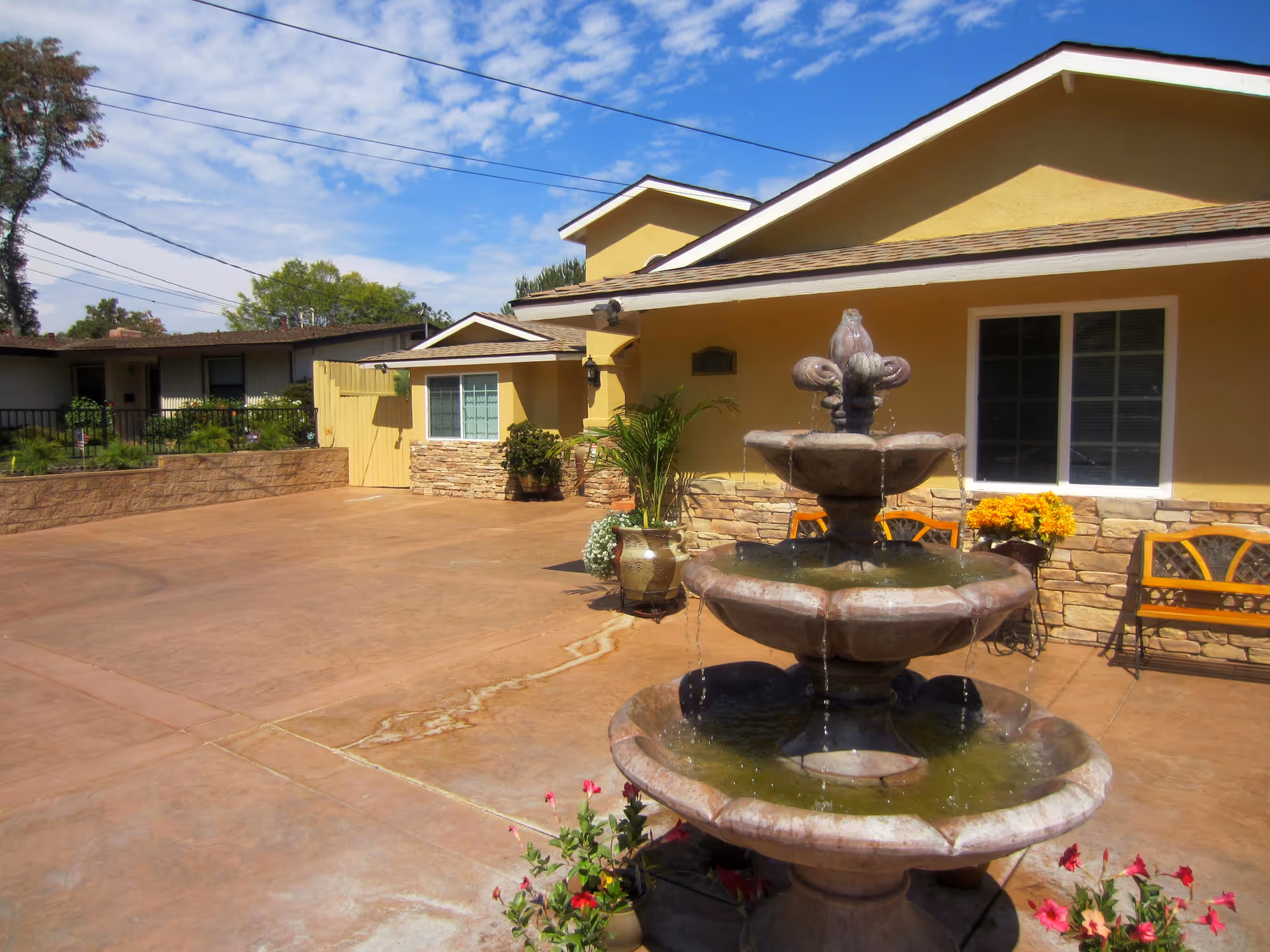 Outdoor courtyard area of a senior living facility with a three-tier stone water fountain in the foreground, potted plants, yellow building with stone accents, benches, and a partly cloudy blue sky.