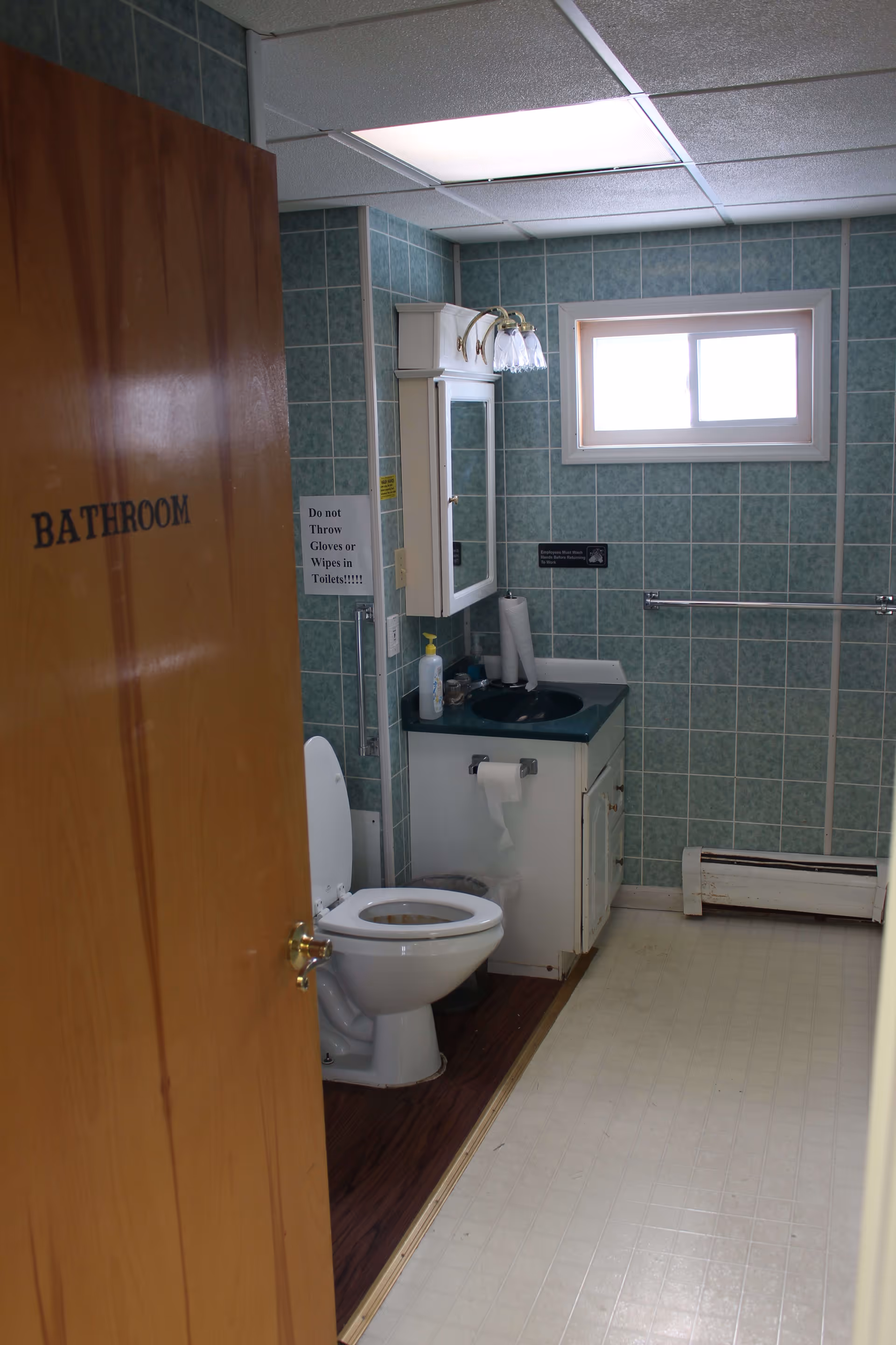 Interior view of a bathroom with green tiled walls, a white toilet with the seat up, a white vanity with a dark green countertop and sink, a medicine cabinet with a mirror above the sink, a window letting in natural light, and a wooden door partially open with the word 'BATHROOM' written on it.