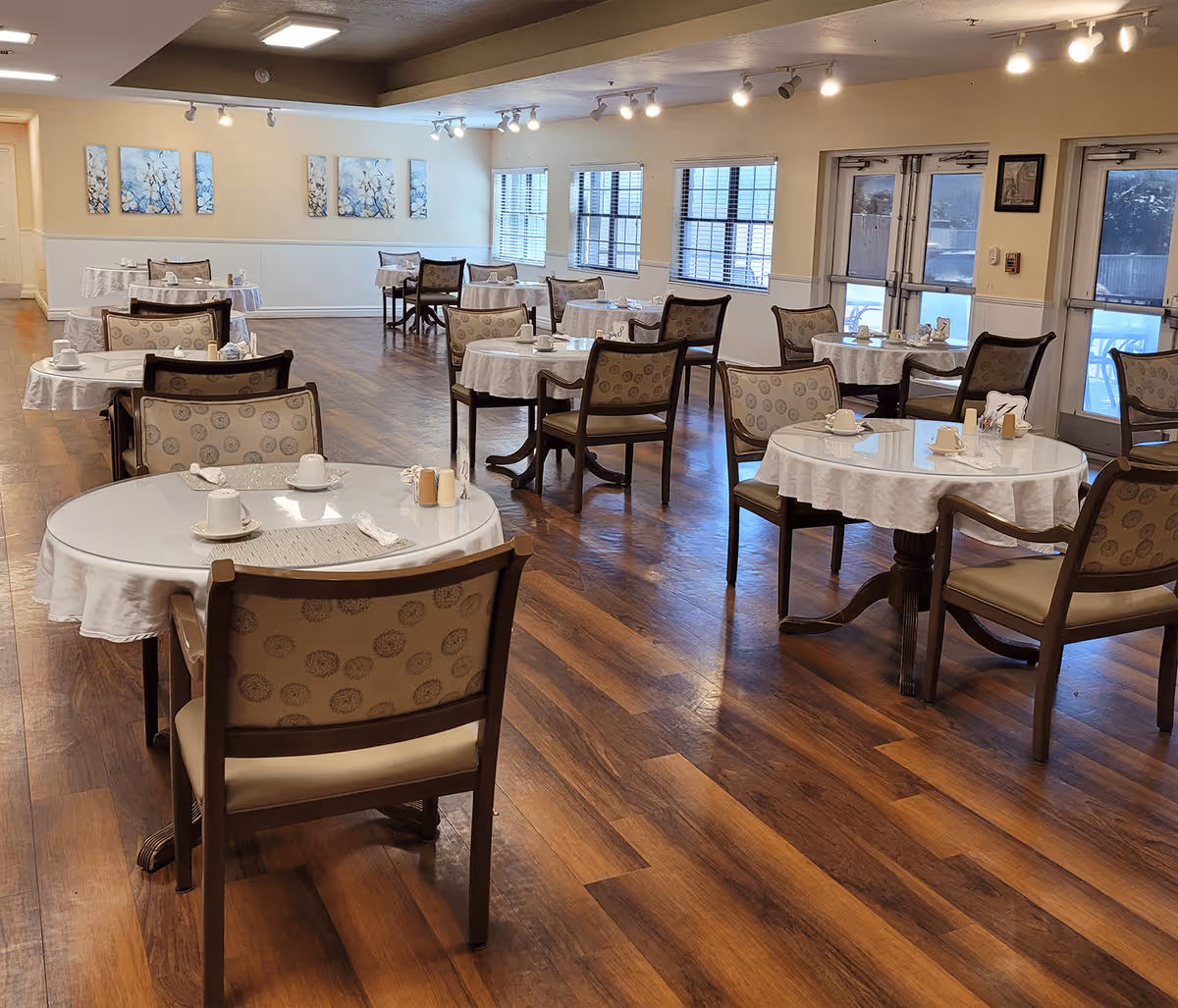 A dining room with multiple round tables covered with white tablecloths, each set with cups, saucers, and napkins. The room has wooden flooring, beige walls with floral artwork, and several windows letting in natural light. Chairs with patterned upholstery surround each table.