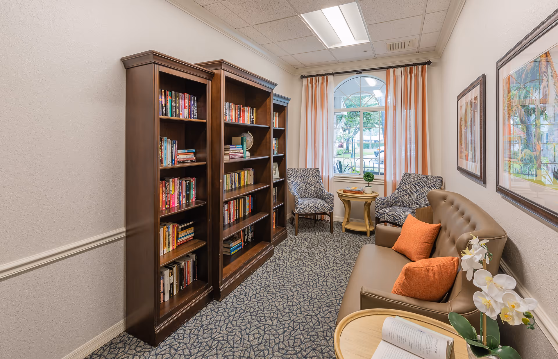 A cozy reading nook in a senior living facility featuring two tall wooden bookshelves filled with books, a brown leather sofa with two orange pillows, two patterned armchairs, a small round wooden table with a plant and books, and a large window with orange and white striped curtains letting in natural light.