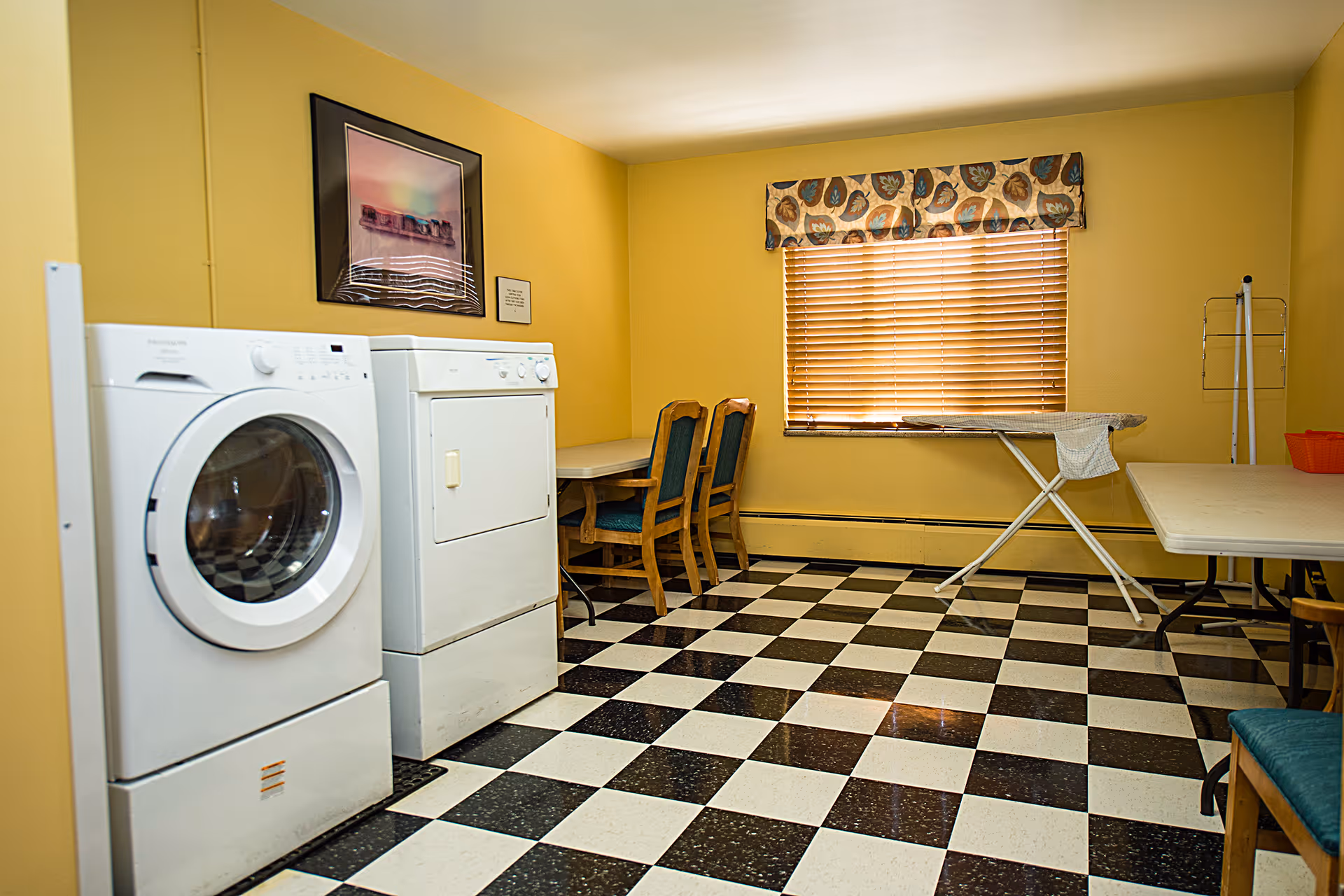 A laundry room with a white front-loading washing machine and a white dryer side by side. The room has yellow walls, a window with wooden blinds and a floral valance, a black and white checkered floor, two wooden chairs with green cushions, a folding table, and an ironing board with a cloth draped over it. A framed picture and a small sign are hung on the wall above the machines.