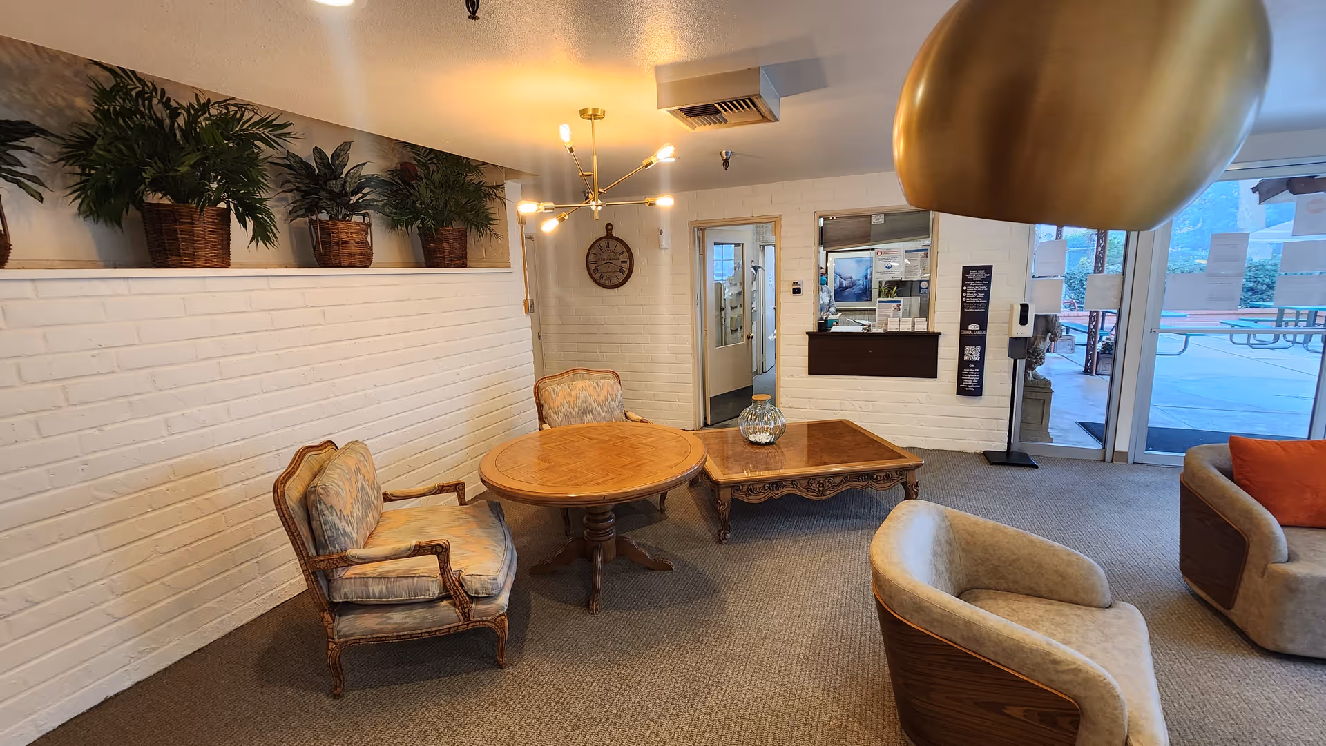 A cozy interior common area with vintage-style armchairs and wooden tables on a carpeted floor. There are potted plants on a ledge along a white brick wall, a modern chandelier hanging from the ceiling, and a clock on the wall. A reception window and glass doors leading outside are visible in the background.
