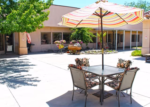 Outdoor patio area at Hearthstone Of Northern Nevada with a table and four chairs under a large striped umbrella. The patio is surrounded by a building with windows and some greenery including trees and potted plants.