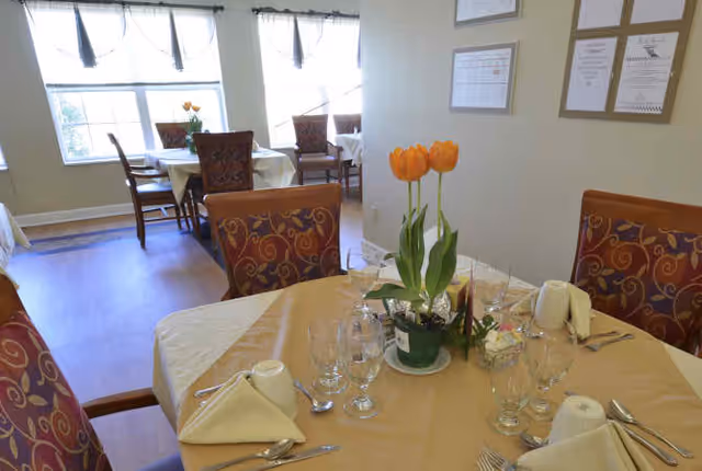 A dining room with tables set for a meal, featuring beige tablecloths, folded napkins, glassware, and silverware. Each table has a centerpiece with orange tulips. The room has large windows with sheer curtains allowing natural light to enter. Framed certificates or documents are hung on the wall.