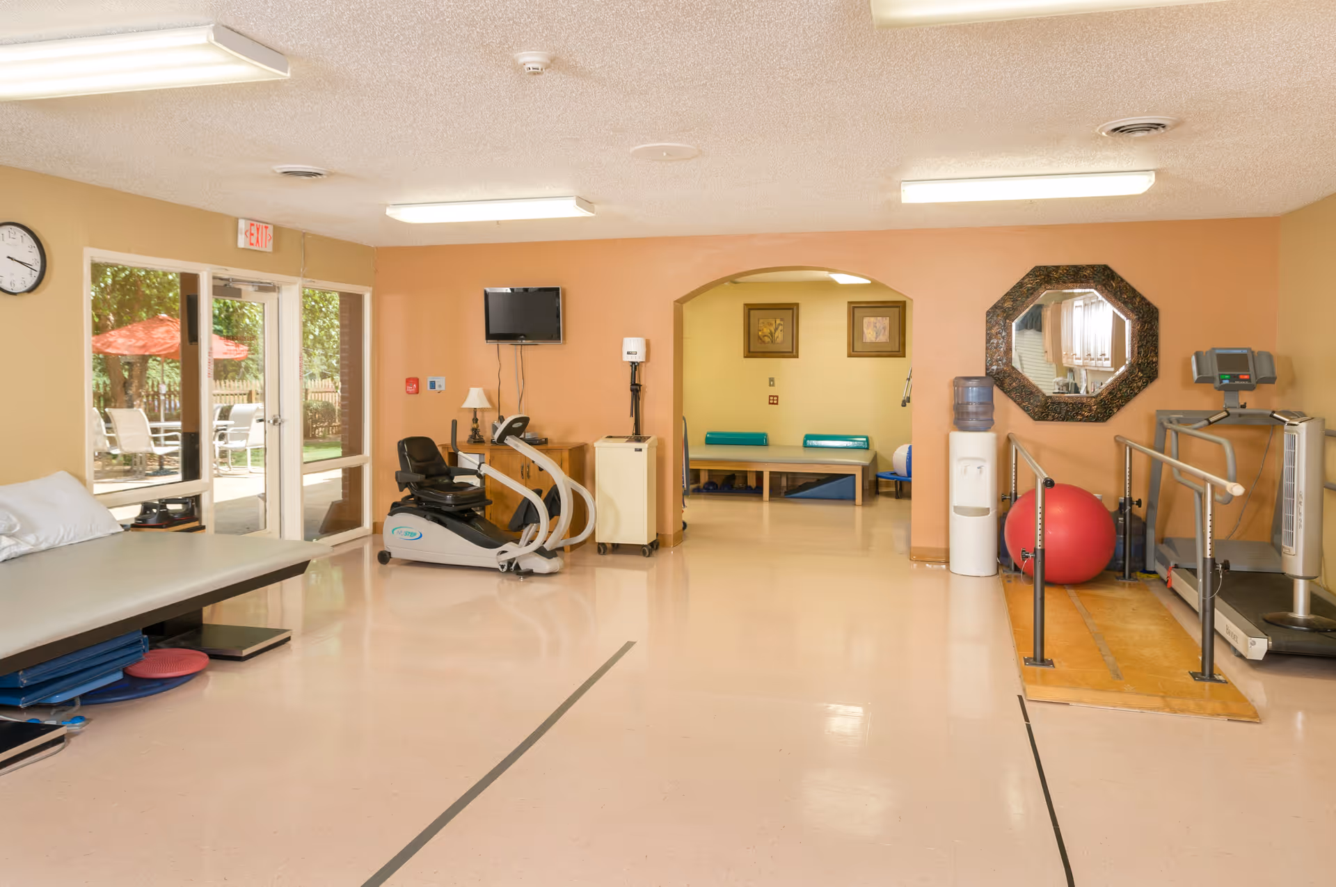A rehabilitation or physical therapy room with exercise equipment including a recumbent bike, treadmill with handrails, a large red exercise ball, and therapy tables. There is a water cooler, a wall-mounted TV, a clock, and glass doors leading outside to a patio area with red umbrellas and chairs.