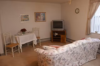 A cozy living room area with a floral patterned sofa facing a small wooden TV stand holding an older model television and a telephone. To the left, there is a small dining table covered with a white tablecloth, set with dishes and a flower centerpiece, accompanied by two chairs. The room has beige walls, two framed pictures hanging above the dining table, a clock on the wall, and a window with beige curtains letting in natural light.