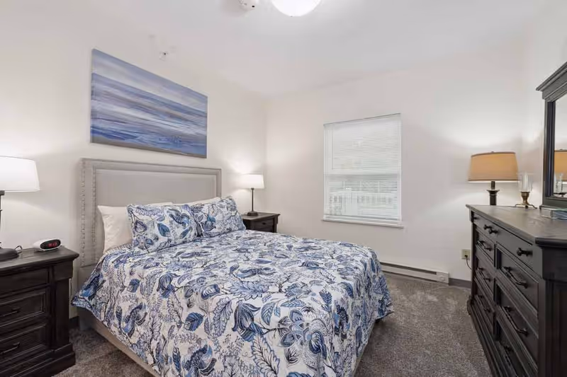 A bedroom with a bed covered in a blue and white floral patterned comforter and matching pillows. The bed has a light gray upholstered headboard. There are two dark wooden nightstands on either side of the bed, each with a lamp. A window with closed blinds is on the wall opposite the bed. A dark wooden dresser with a mirror and a lamp is on the right side of the room. The walls are white and the floor is carpeted in gray.