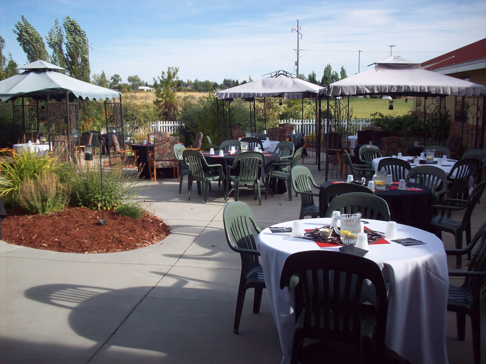 Outdoor patio with round tables set for dining, green plastic chairs, and canopied gazebos.