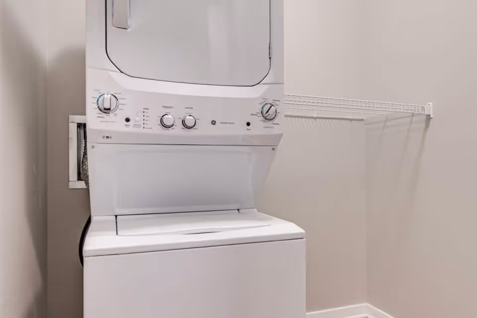 A small laundry area with a stacked white washer and dryer unit against a beige wall. There is a white wire shelf mounted on the right wall above the washer and dryer.