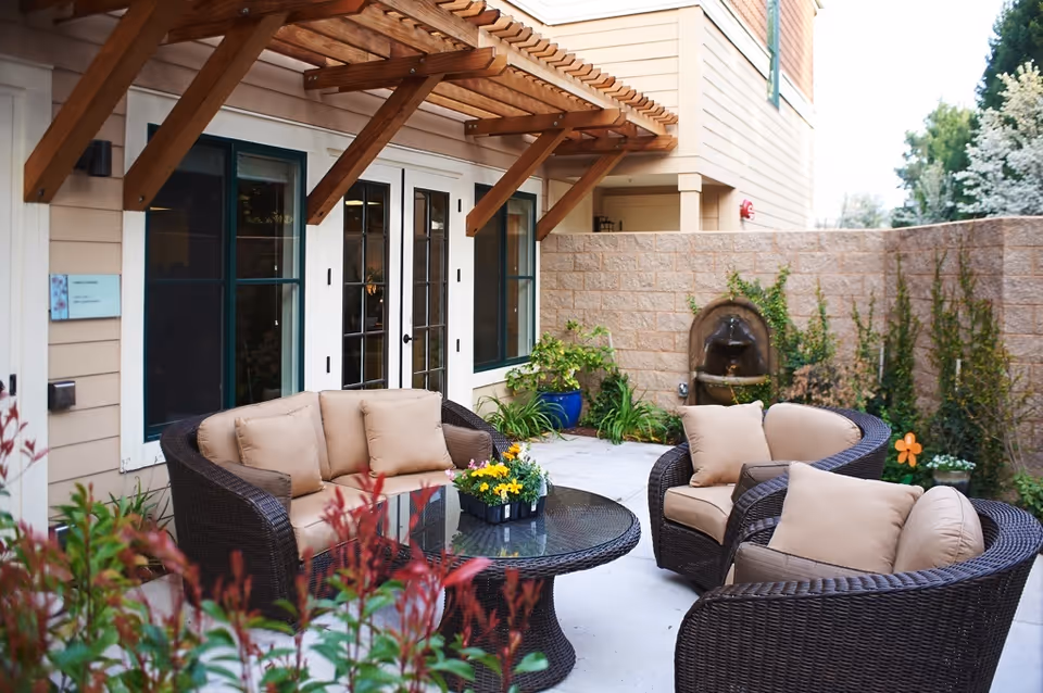 Outdoor patio area with wicker furniture including a sofa and two armchairs with beige cushions arranged around a round glass-top table with a flower centerpiece. The patio is adjacent to a building with beige siding and white-framed windows and doors. There is a wooden pergola overhead and a stone wall with a decorative water fountain and plants in the background.