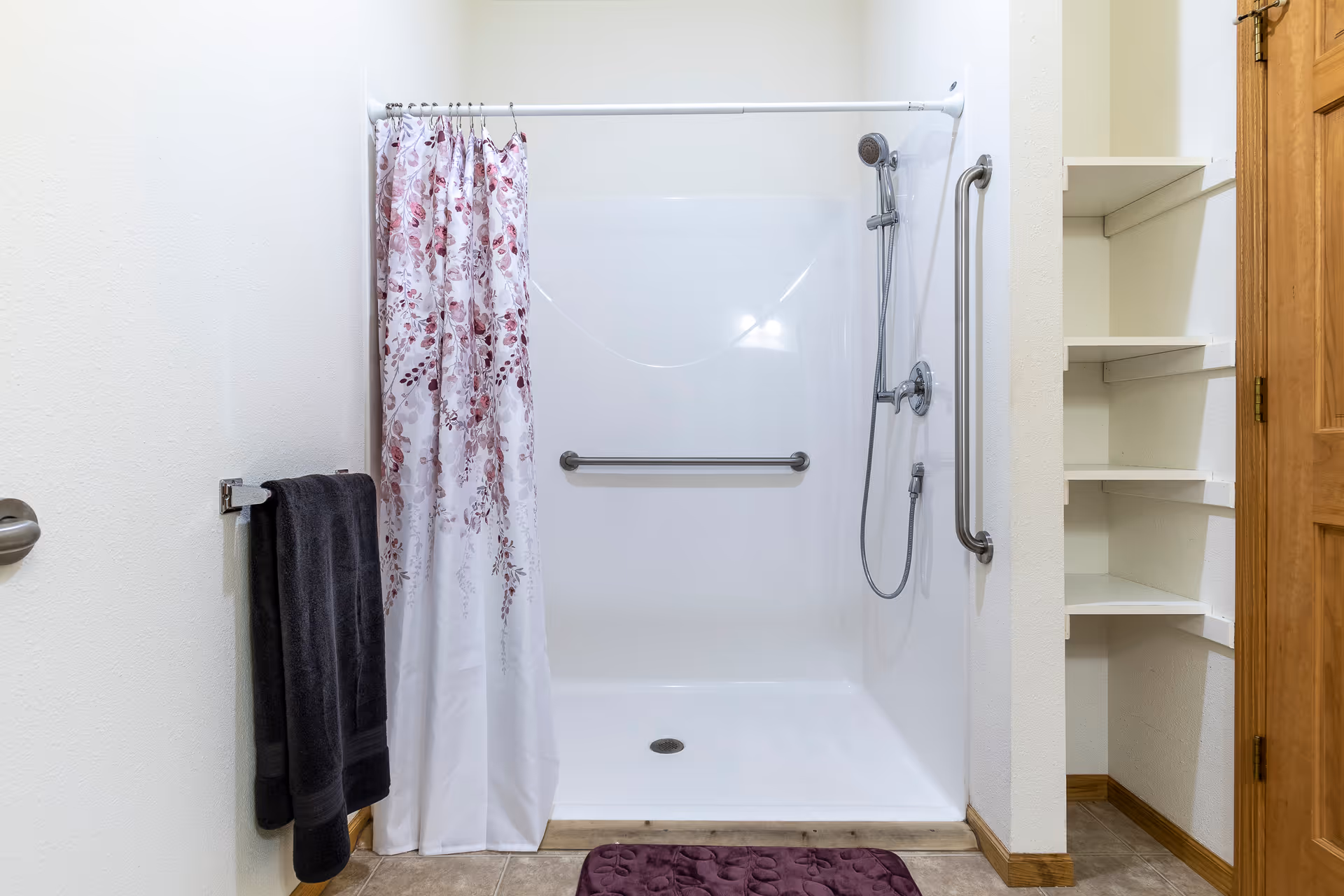 Walk-in shower with a floral curtain, grab bars, handheld showerhead, towel on a rack and adjacent shelving.
