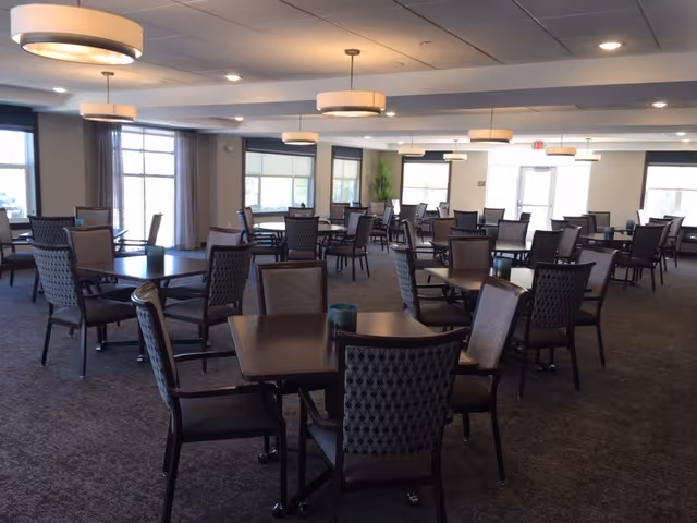 Spacious dining room with multiple dark wood tables and upholstered chairs, pendant lights, and large windows.