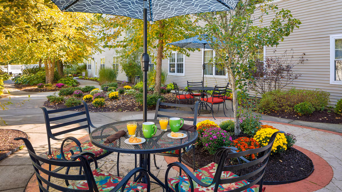 Outdoor patio area at a senior living facility with a round metal table and four chairs with floral cushions. The table has two green mugs, two glasses of orange juice, and two plates with pastries. There are large umbrellas providing shade, colorful flower beds, trees, and a beige building with windows in the background.