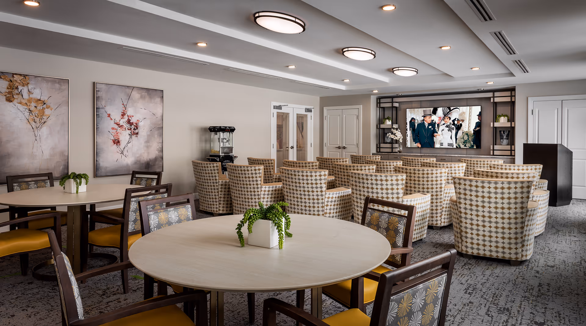 A well-lit common area with round tables and chairs in the foreground and rows of patterned armchairs facing a large flat-screen TV mounted on the wall. The room features modern ceiling lights, two abstract paintings on the wall, a popcorn machine, and a podium near the TV.