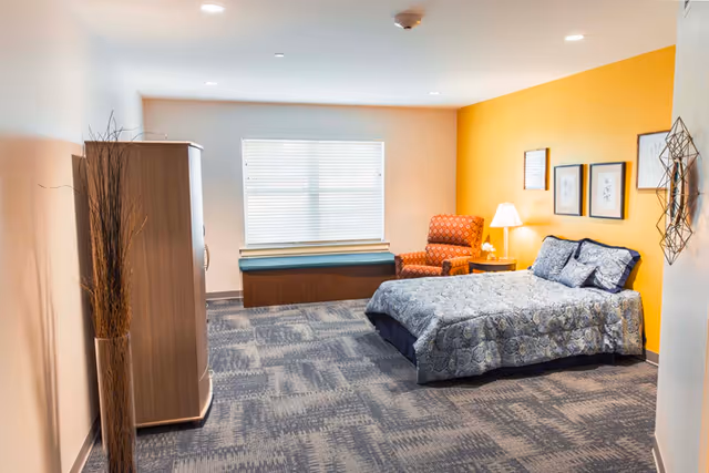 A bright bedroom with a bed, nightstand and lamp, patterned armchair, wardrobe and window bench against a yellow accent wall.