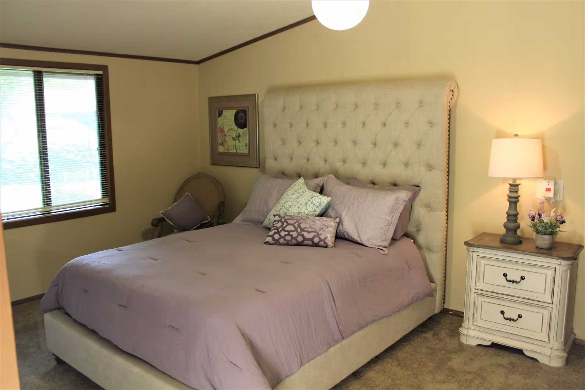 A bedroom with a large tufted beige headboard and a bed made with light purple bedding and decorative pillows. There is a window with blinds on the left wall, a framed floral picture above a chair with a purple cushion in the corner, and a white nightstand with two drawers on the right side of the bed. A lamp and a small flower pot are on the nightstand.