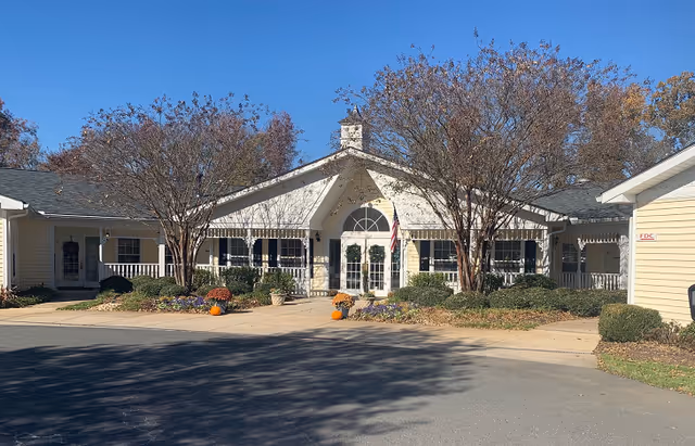Entrance of a single-story senior living building with a covered porch, American flag, landscaping and pumpkins under a clear blue sky.