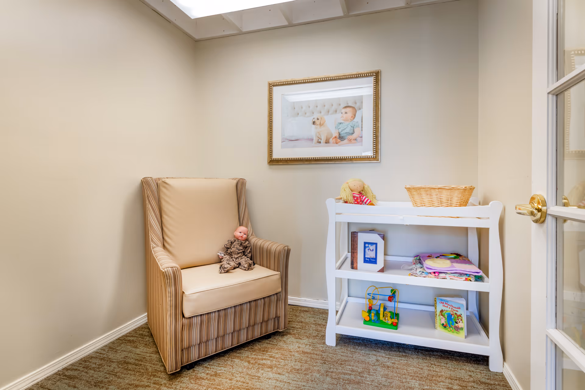 Small interior room with a striped armchair holding a doll, a white shelving unit with toys and a basket, and a framed picture on the wall.