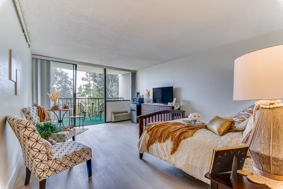 A bright and cozy senior living bedroom with a bed covered in a beige quilt and brown throw blanket, a wooden dresser with a TV on top, two patterned chairs with a small table between them, and a sliding glass door leading to a balcony with outdoor seating and greenery visible outside.