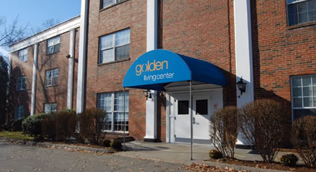 Exterior view of a brick building with multiple windows and a blue awning above the entrance that reads 'golden living center'. There are trimmed bushes along the front of the building and a paved area in front.