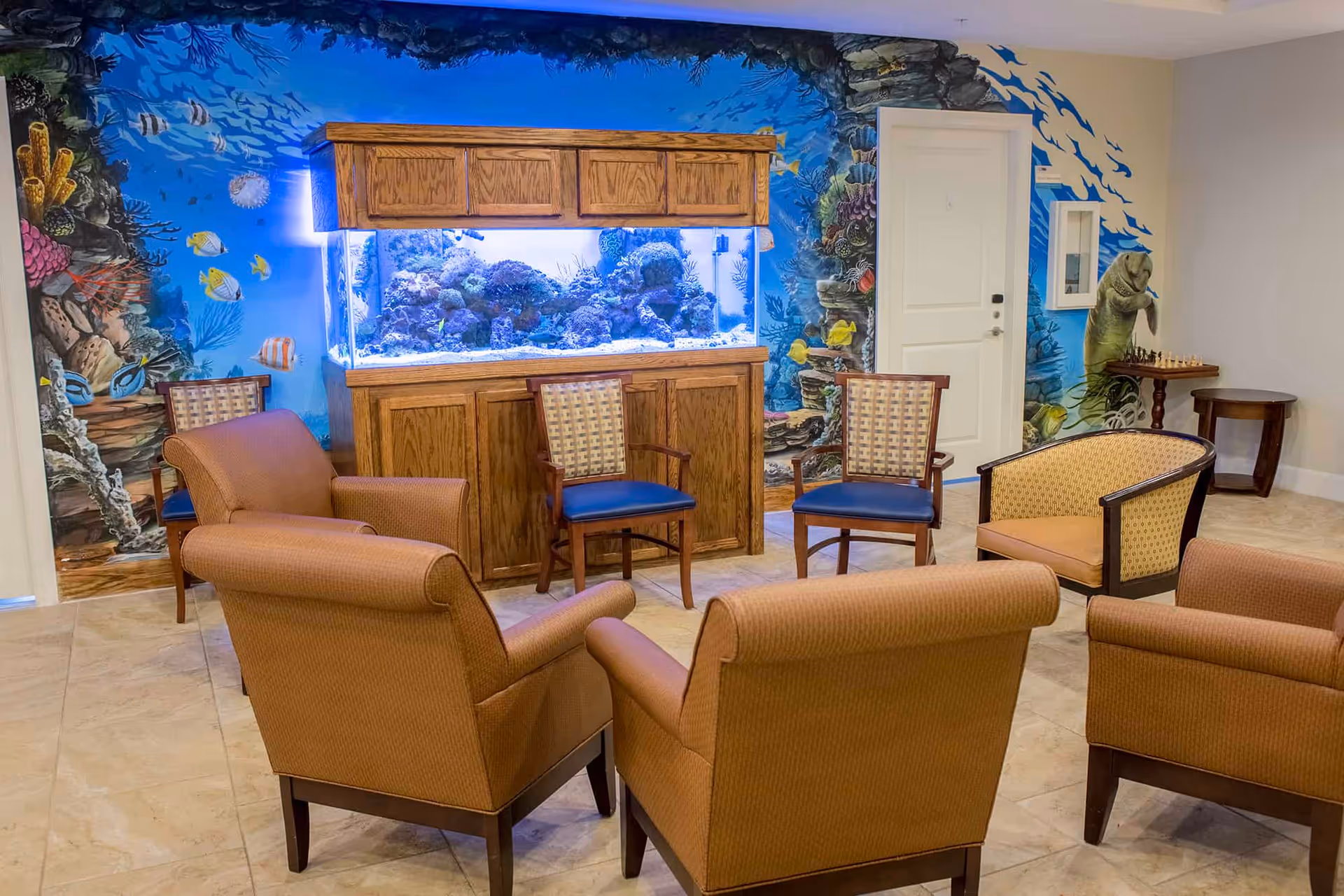 A cozy seating area with several upholstered armchairs and wooden chairs arranged around a large aquarium filled with coral and fish. The wall behind the aquarium features a colorful underwater mural with various fish and sea creatures. A small table with a chessboard and a manatee sculpture is visible near a white door.