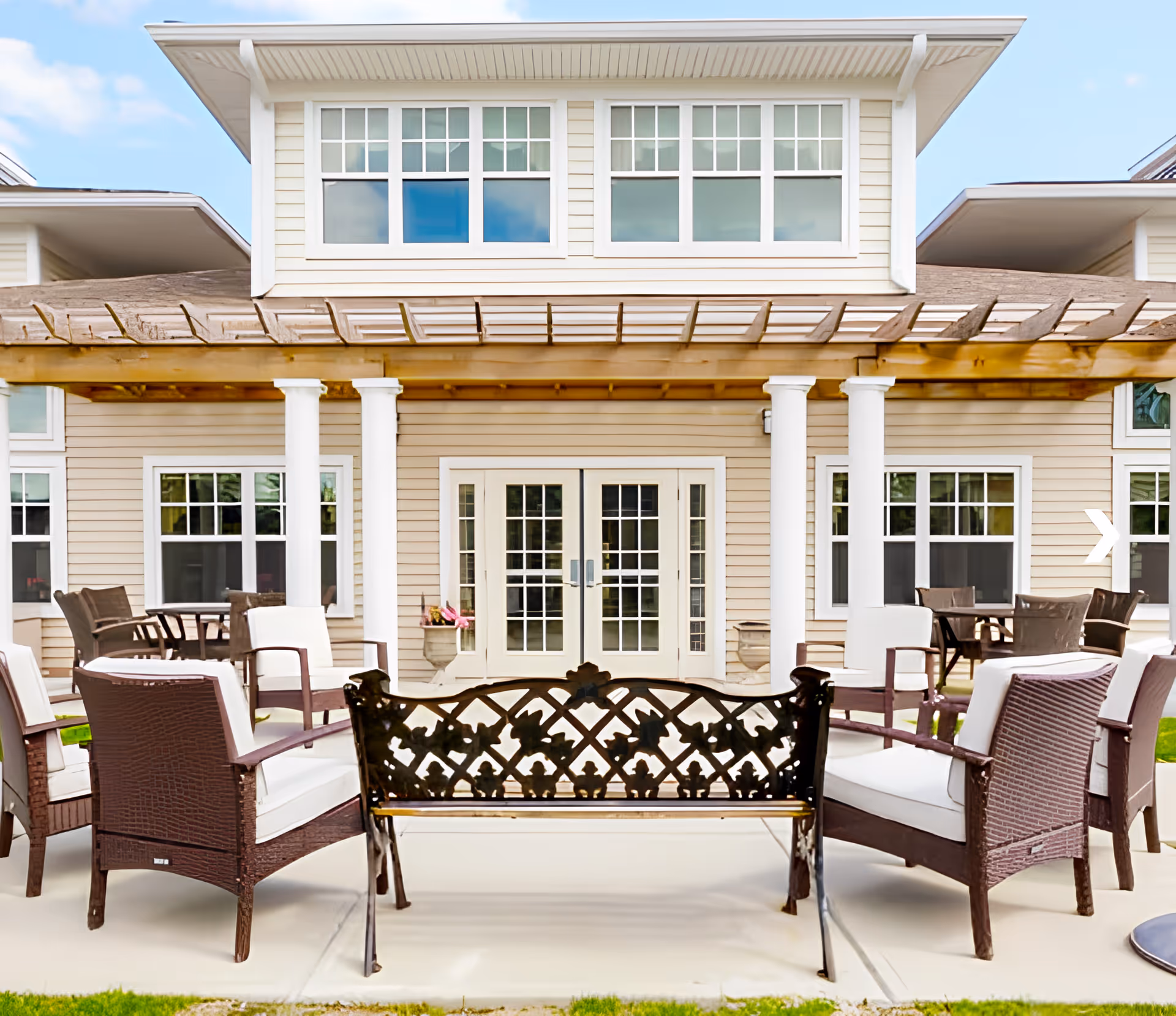 Outdoor patio seating with wicker chairs and a decorative metal bench in front of a two-story building with columns and a pergola.