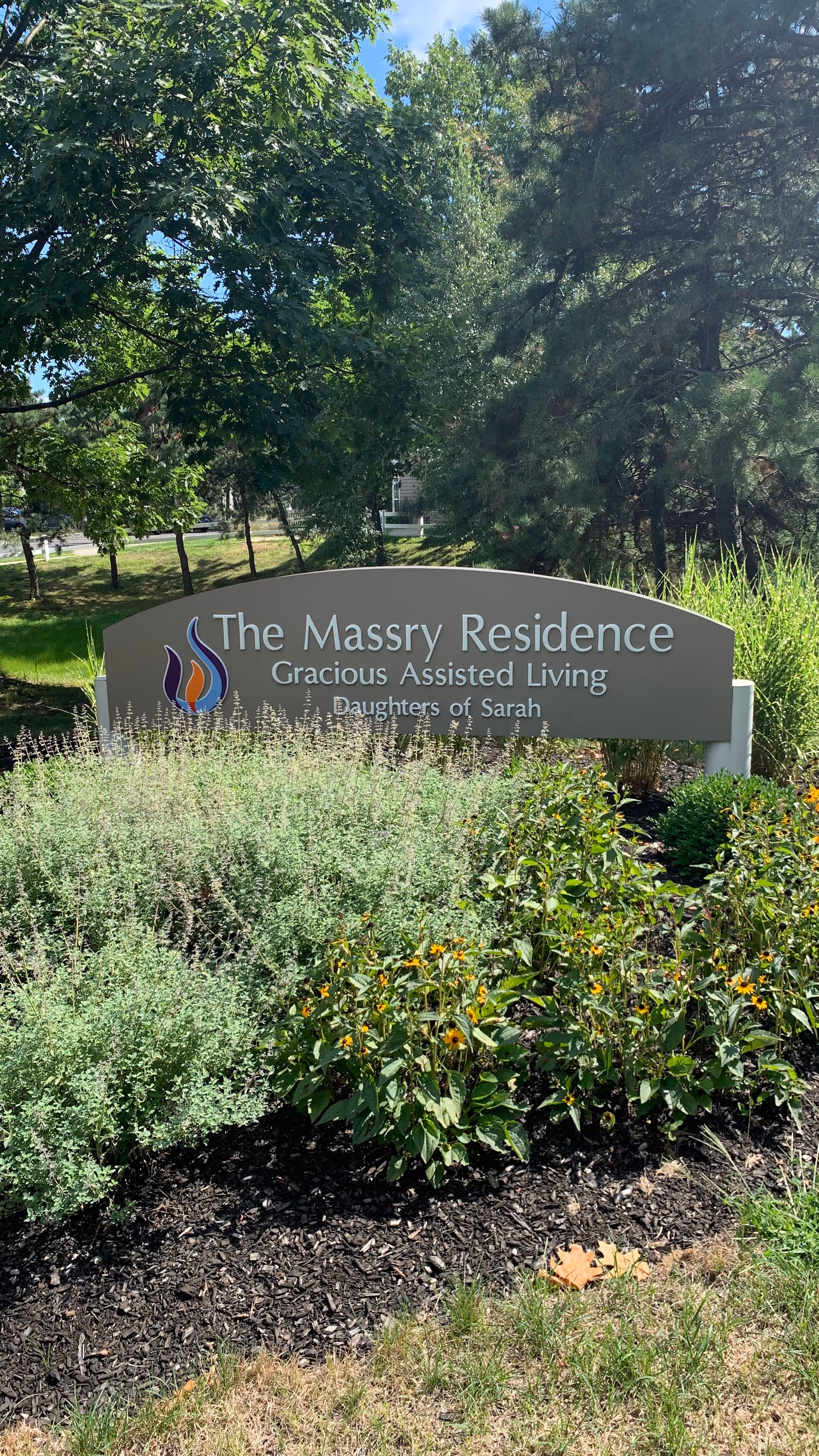 Outdoor sign for The Massry Residence Gracious Assisted Living, Daughters of Sarah, surrounded by green plants and flowers with trees in the background.