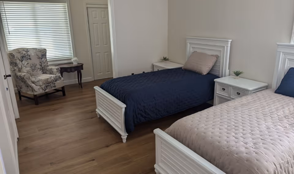 A bedroom with two single beds, one with a navy blue quilt and the other with a light beige quilt. Each bed has a white wooden headboard and a matching white nightstand with small plants on top. There is a floral-patterned armchair near a window with closed blinds and a small wooden table beside it. The room has light-colored walls and wooden flooring.