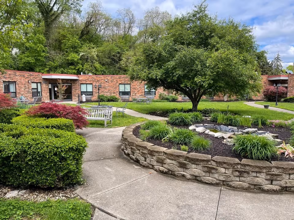 Brick single-story building surrounding a landscaped courtyard with a central tree, benches, walkways, and planting beds.