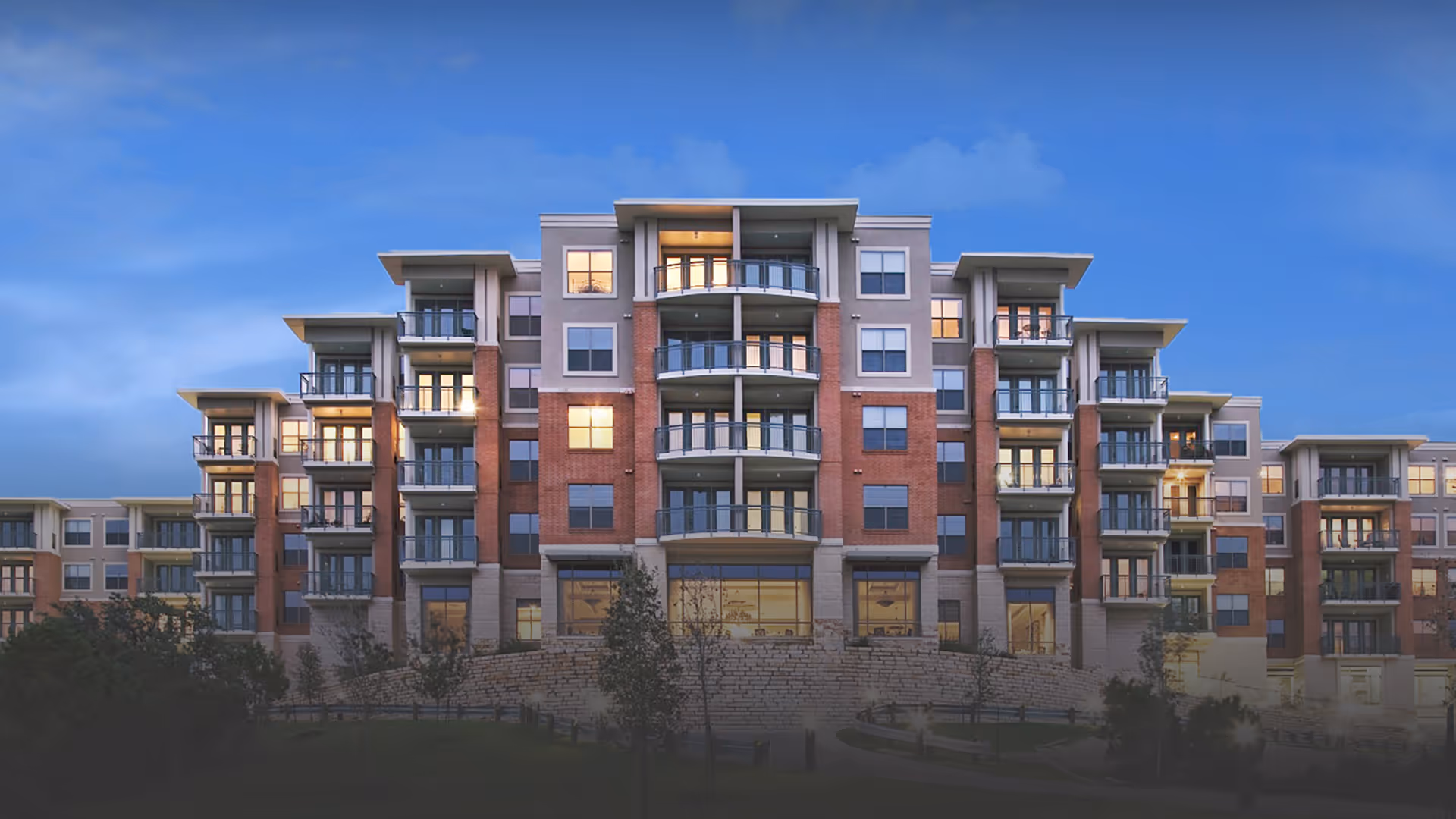 Exterior view of a multi-story senior living facility building with balconies and large windows, illuminated from inside during dusk with a clear blue sky in the background.