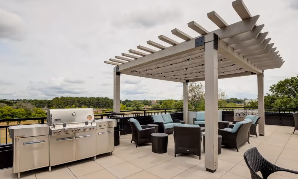 Rooftop patio with a pergola, wicker seating and cushions, and a stainless steel grill overlooking trees.