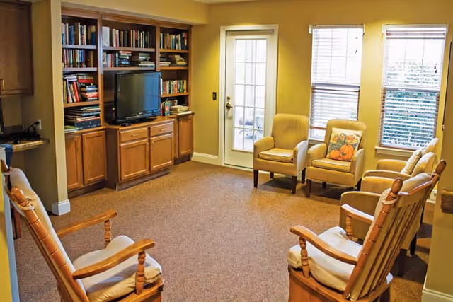A cozy living room with beige walls and carpet, featuring a wooden built-in bookshelf and cabinet unit with a television. The room has several cushioned chairs arranged in a circle, including two wooden rocking chairs and three upholstered armchairs near a door with glass panes and two windows with blinds.