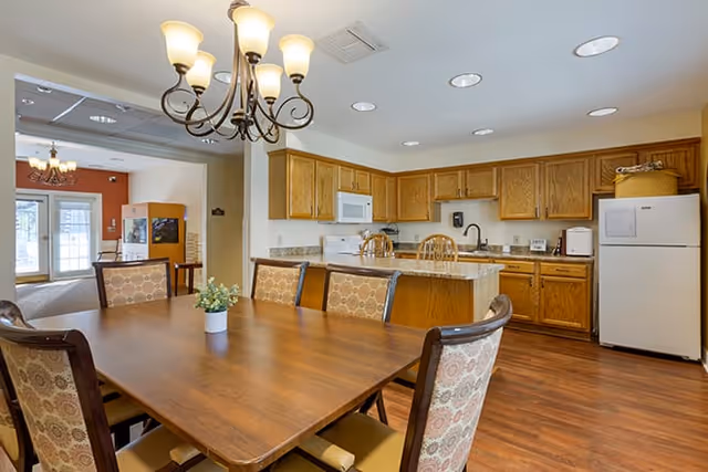 Interior view of a senior living facility dining area and kitchen with a wooden dining table surrounded by six upholstered chairs, a chandelier overhead, wooden cabinetry, a white refrigerator, microwave, and kitchen island with two wooden bar stools.