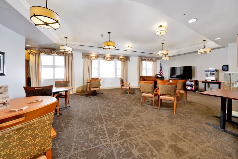 Spacious senior living common room with tables and chairs, pendant lights, a TV, and large windows with drapes.